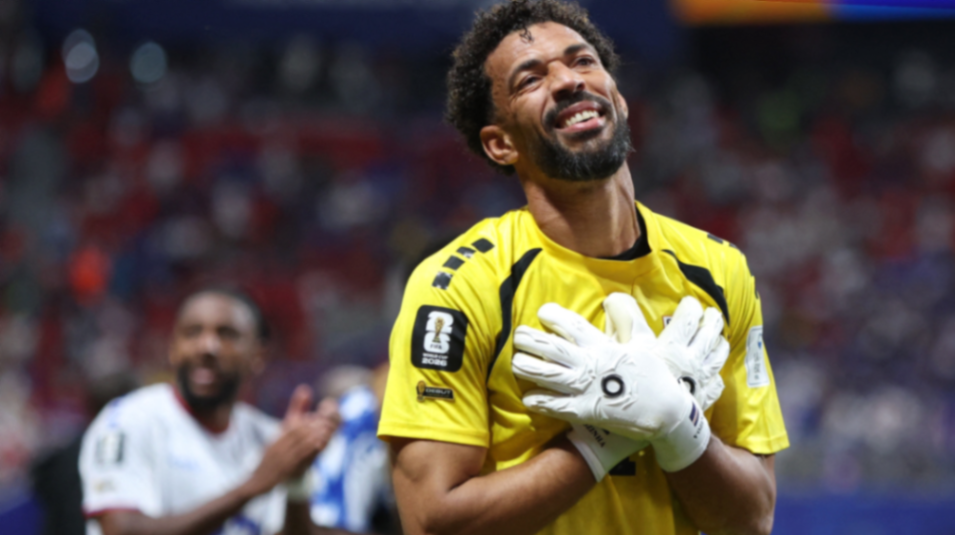 Goleiro em uniforme amarelo sorri e faz gesto de gratidão, enquanto um companheiro aplaude ao fundo em estádio.